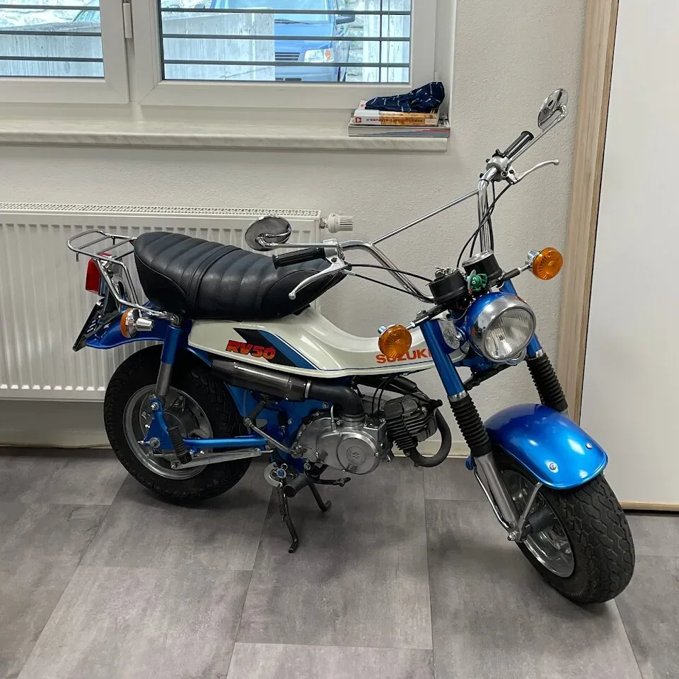 Kleines blaues Suzuki-Motorrad, Modell RV90, steht in einem Raum mit Fenster im Hintergrund.